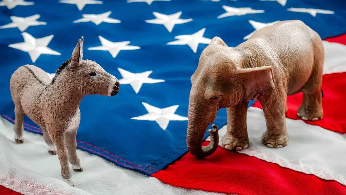 Two plastic figurines, a donkey and an elephant, stand facing each other on a folded American flag with stars and stripes…