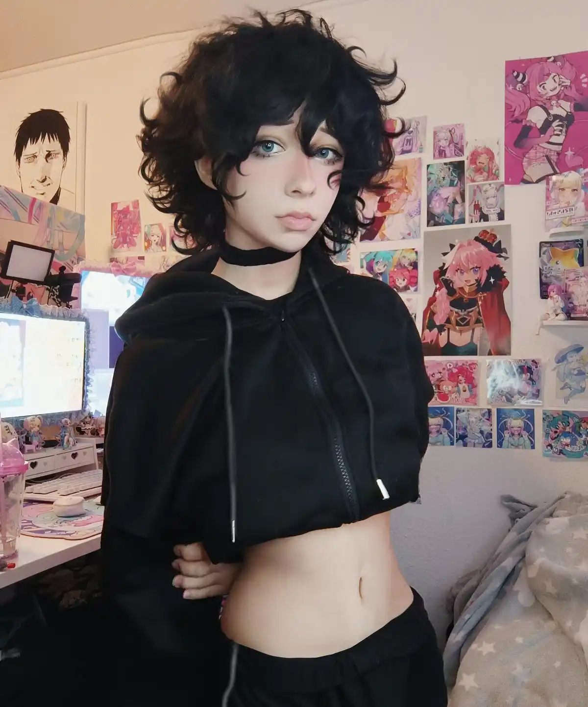 A person with short black curly hair and blue eyes wears a cropped black hoodie and black choker, standing in a bedroom…