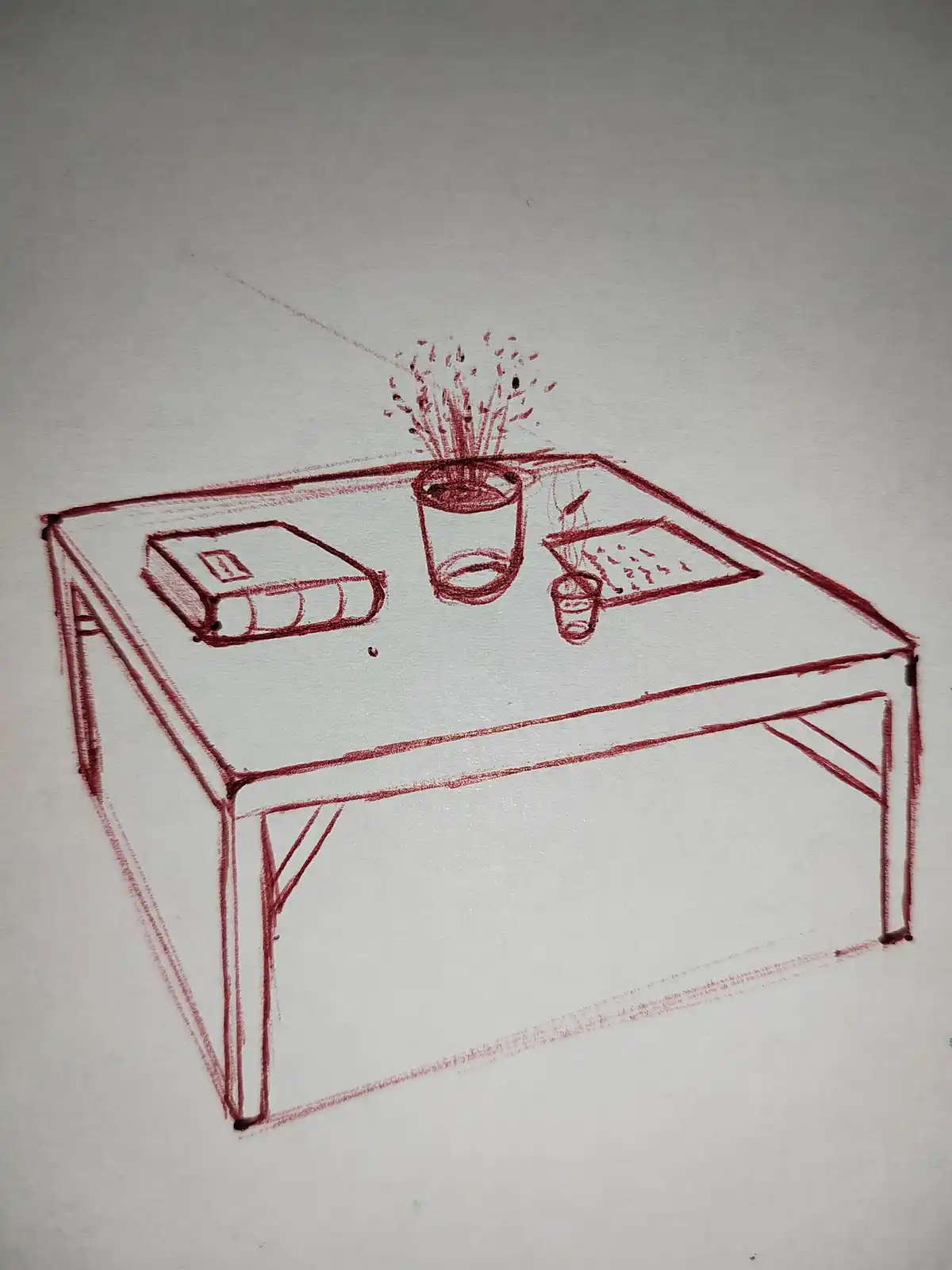 A red crayon drawing on white paper shows a table with a book, a glass of liquid exploding upwards, and a small cup. The…