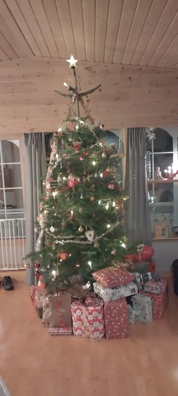 a full, decorated christmas tree with lights, ornaments, and a star topper stands in a room with wooden floors and ceilings…