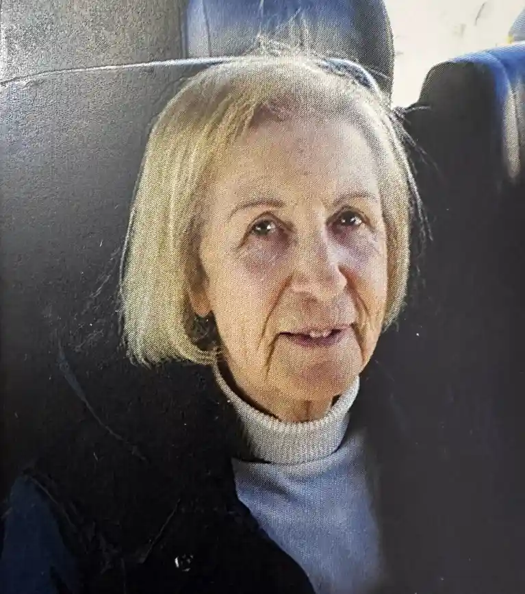 An older woman with short blonde hair, wearing a light blue turtleneck under a dark jacket, sits in a dark blue upholstered…