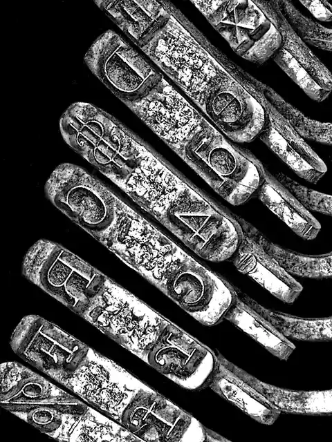A black-and-white image shows a diagonal row of worn, metallic letterpress printing blocks, each engraved with a single…