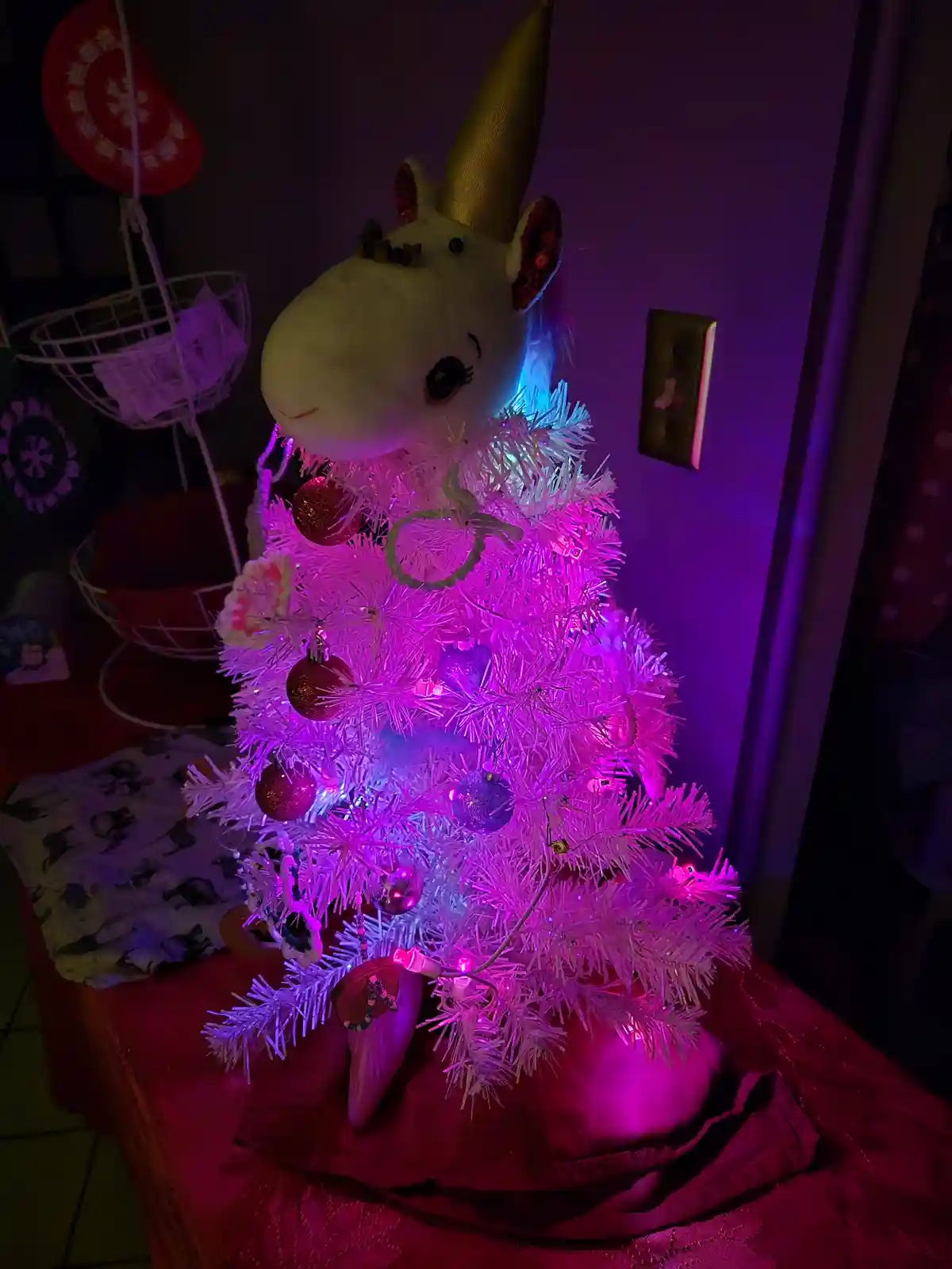 A small white artificial Christmas tree is decorated with pink and purple lights, red and purple ornaments, and topped with…