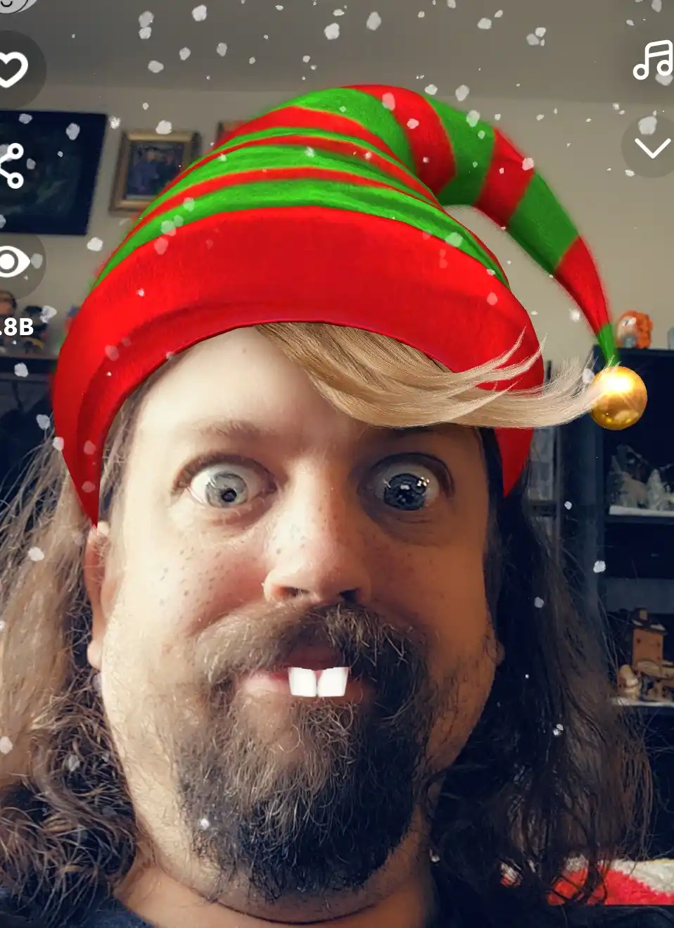 A man with a beard and long hair wears a red and green striped elf hat with a gold bell, his eyes are wide open and his…