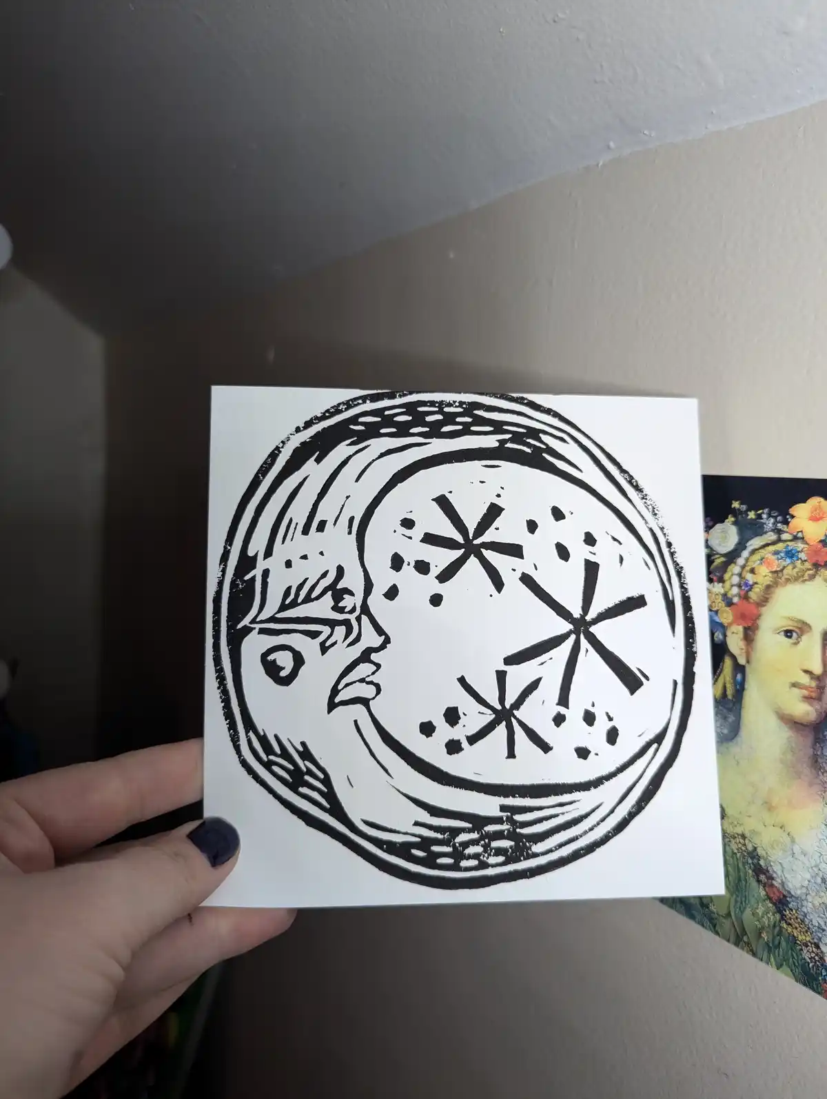 A hand holds a square white card with a black ink drawing of a crescent moon containing a stylized human face profile and…