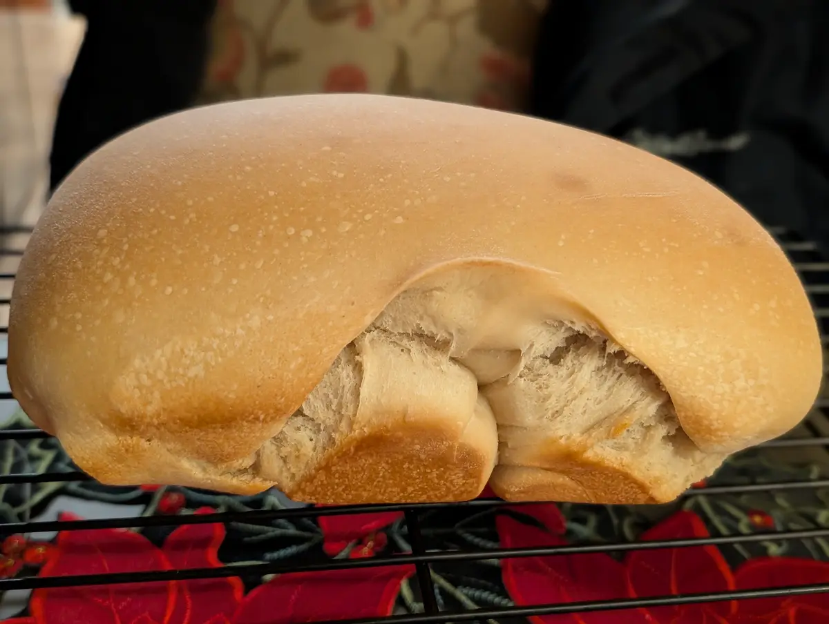 A close-up of a golden-brown, soft bread roll resting on a cooling rack, with its top slightly split open to reveal the…