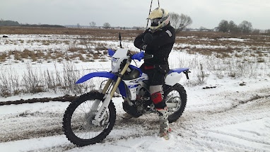 A person in full motocross gear, including helmet and gloves, stands beside a blue and white dirt bike in a snowy field. The…