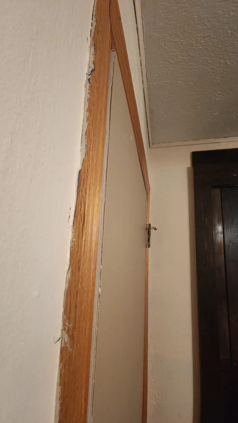 A close-up photo shows a warped, uneven wooden door frame with peeling paint and a small brass hinge, set against textured…