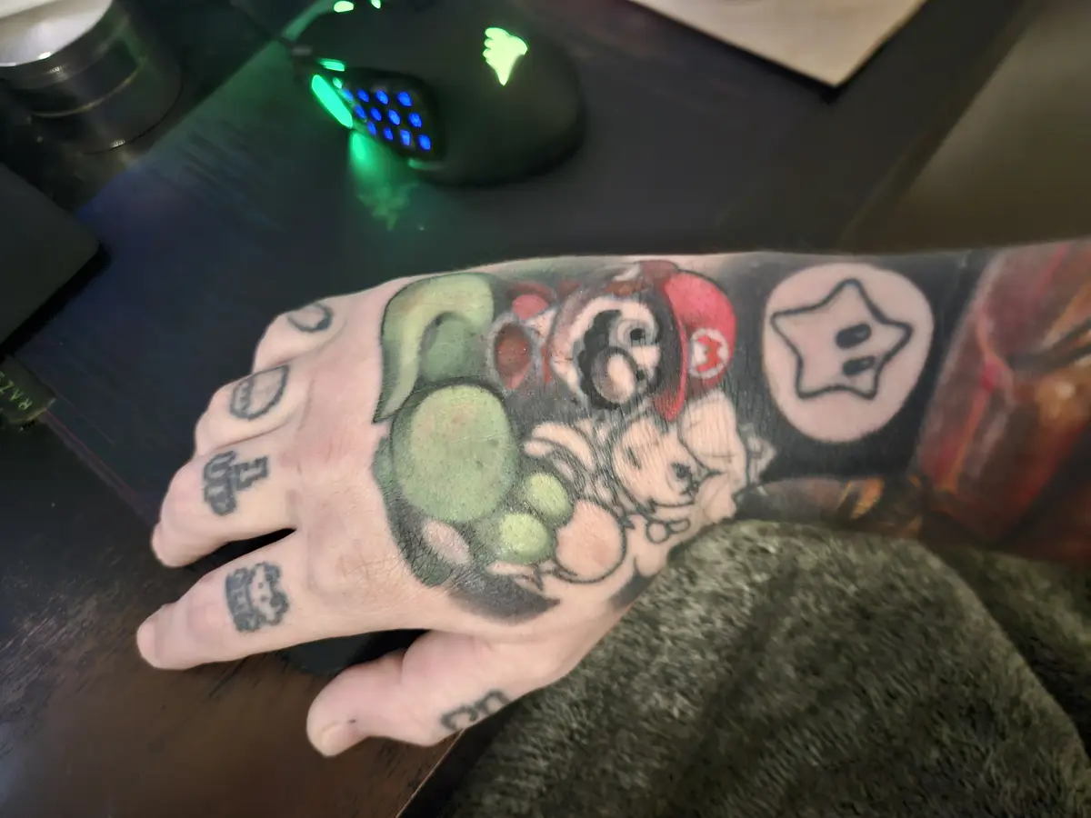 A close-up photo shows a person's left hand and wrist covered in colorful Super Mario-themed tattoos, including Luigi…