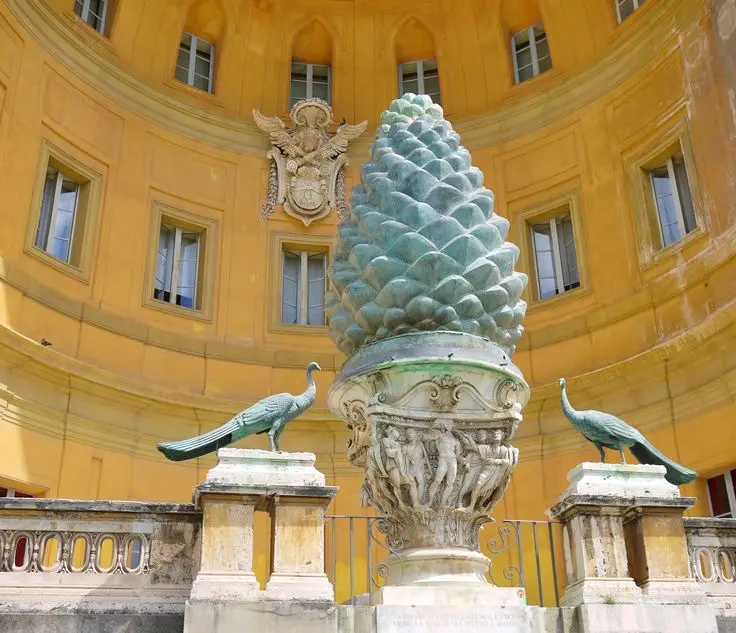 A large, ornate bronze pinecone sculpture stands on a pedestal in front of a yellow, circular building with arched windows…