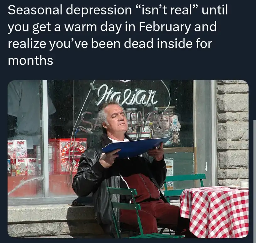 A man in a black leather jacket and red pants sits on a green chair outside a storefront with 'Italian Sausage' written in…