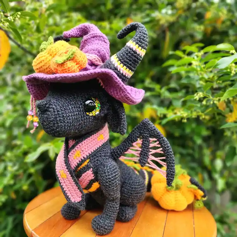 A crocheted black dragon plushie sits on a wooden table outdoors, wearing a purple witch's hat adorned with a crocheted…
