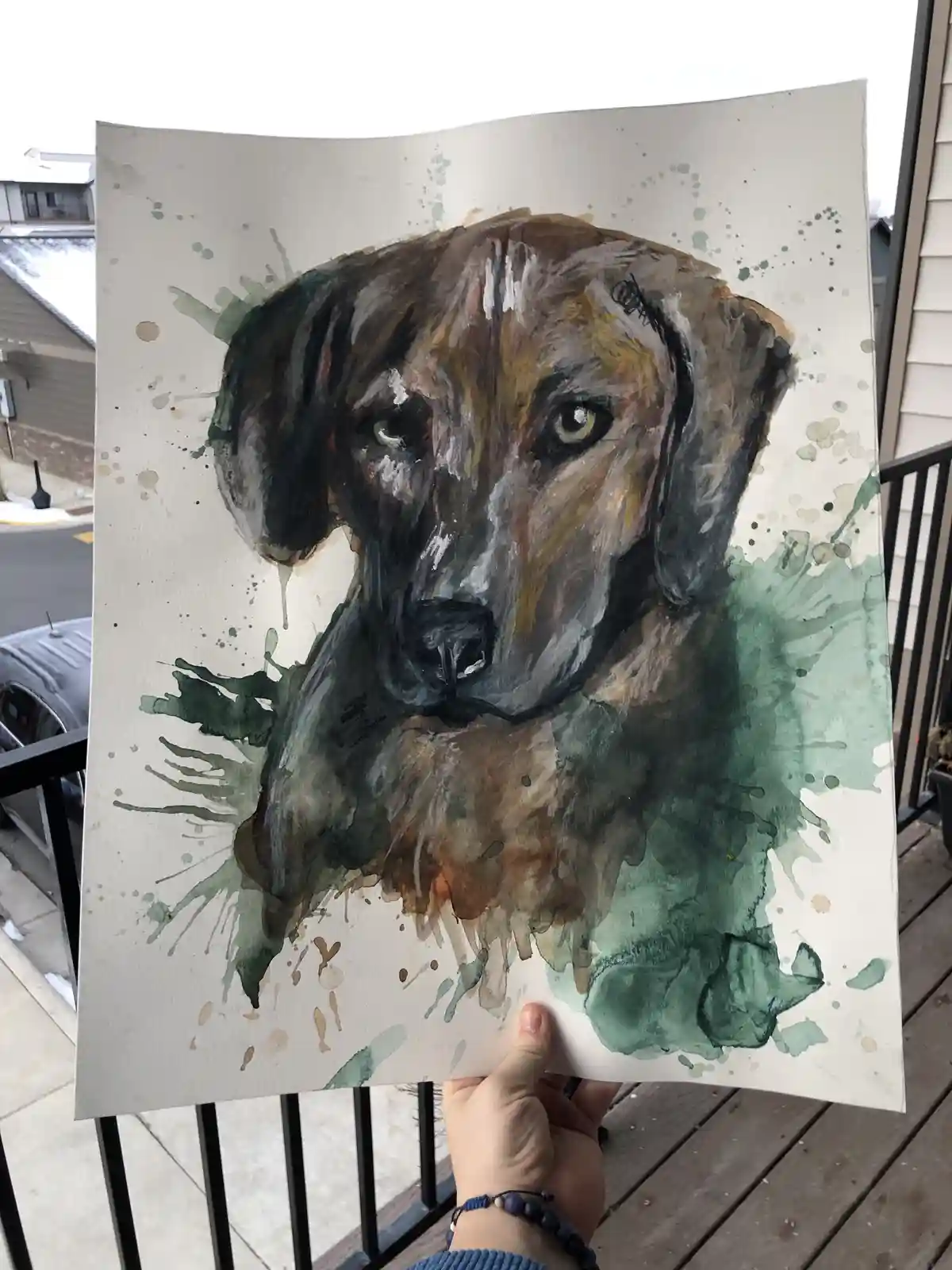 A hand holds up a watercolor painting of a dog's head, featuring brown, black, and green splatters around the portrait. The…