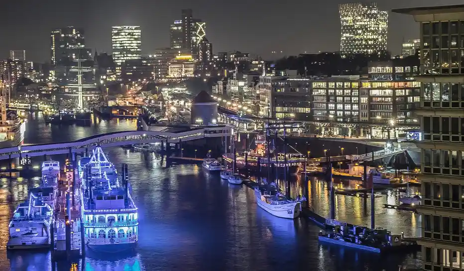 A nighttime city harbor scene with illuminated modern buildings, boats docked along the water, and a bridge spanning the…