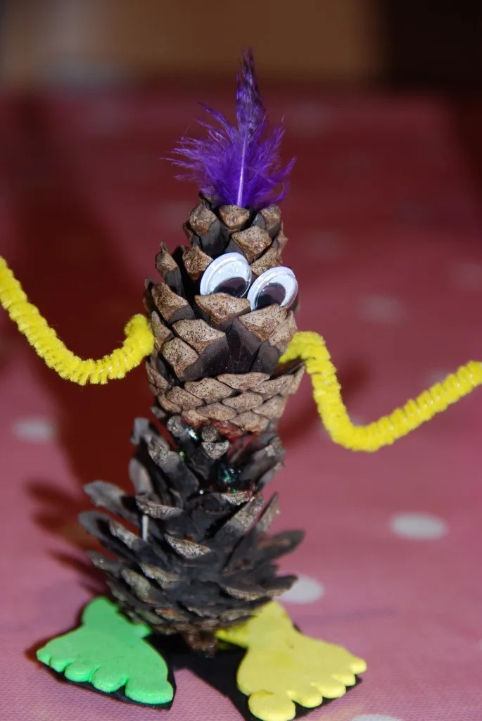 A handmade craft figure made from a pinecone, decorated with googly eyes, a purple feather on top, yellow pipe cleaner arms…