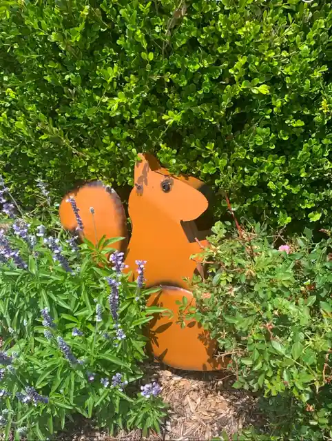 An orange metal squirrel garden ornament is partially obscured by green bushes and purple lavender flowers, with the…