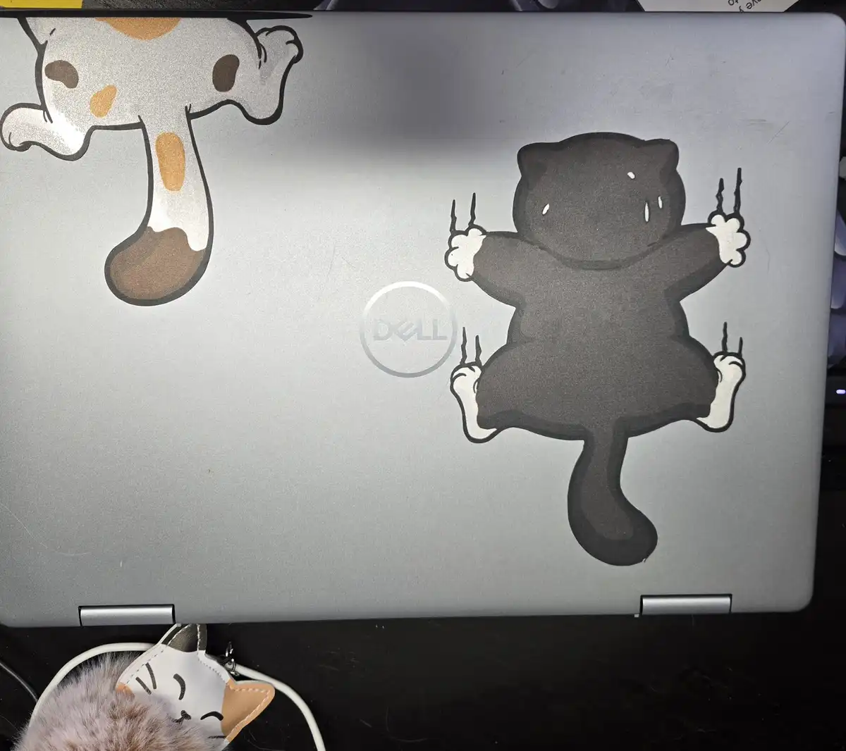 The image shows the back of a silver Dell laptop with two cartoon cat stickers. One is a white cat with brown spots on the…
