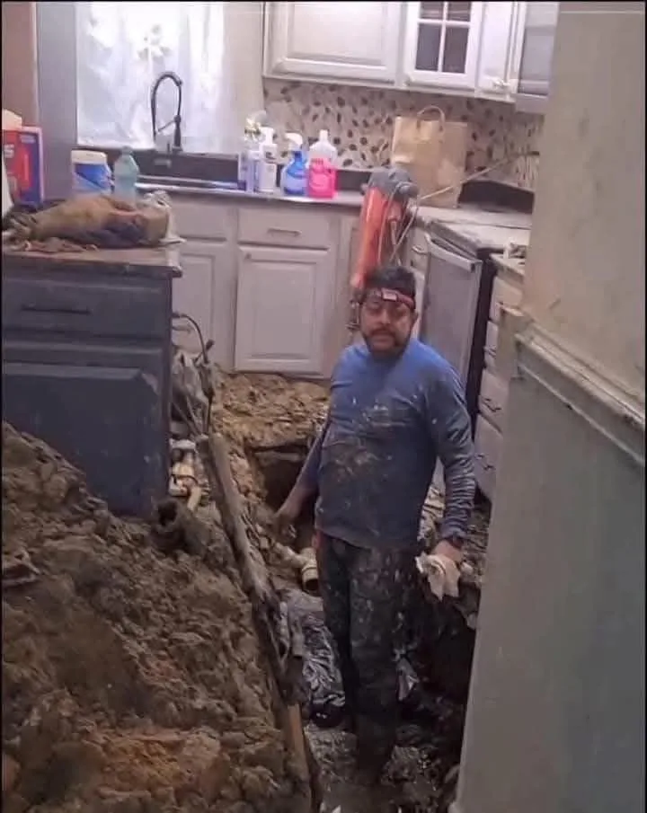 A man covered in mud and grime stands in a kitchen with a large trench dug through the floor, surrounded by dirt and…