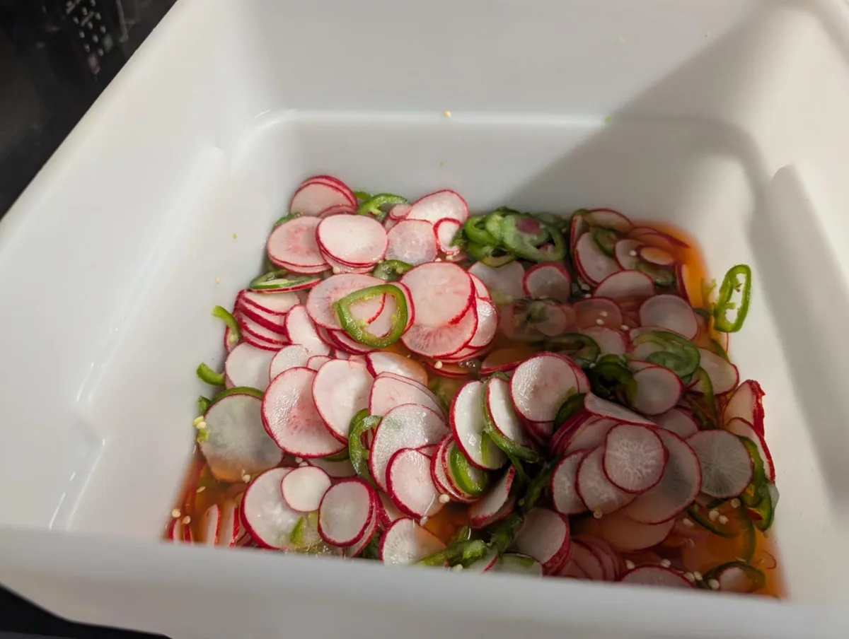 A white rectangular container holds thinly sliced radishes with red skins and white interiors, mixed with sliced green…