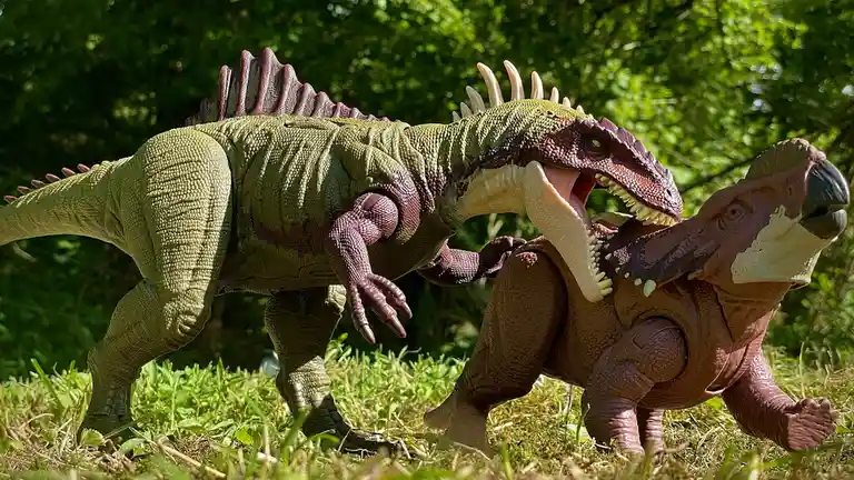 Two large, detailed plastic dinosaur toys are posed in a dynamic, aggressive combat stance on green grass with trees in the…