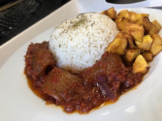 a close-up photo of a white plate with a mound of white rice, a portion of meat in a red sauce, and fried plantains. The…
