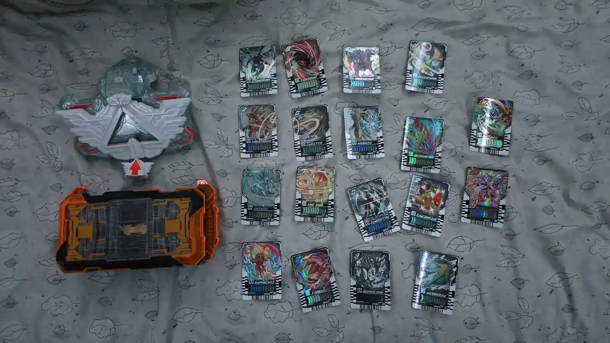 An overhead shot displays a collection of colorful, collectible Kamen Rider card game cards arranged in rows on a grey…