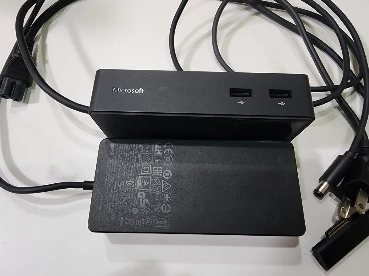 A black Microsoft-branded USB hub sits atop its matching power adapter, both resting on a white surface with tangled black…
