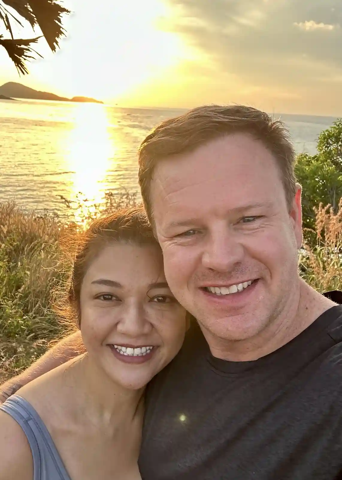 A smiling man and woman pose closely for a selfie against a golden sunset over the ocean, with palm fronds visible in the…