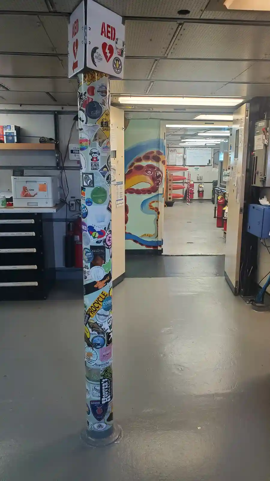 A tall, cylindrical column in an indoor space, heavily covered with colorful stickers from various brands and events, has a…