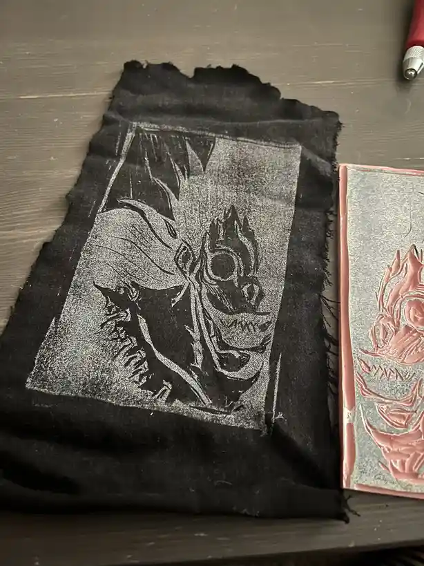 A black, frayed piece of fabric with a faded, white-inked graphic of a demonic, spiky-haired character with a skull-like…