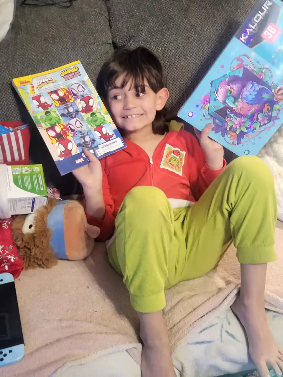 A young child in red and green pajamas sits on a couch holding two colorful activity books — one Marvel Spider-Man themed…