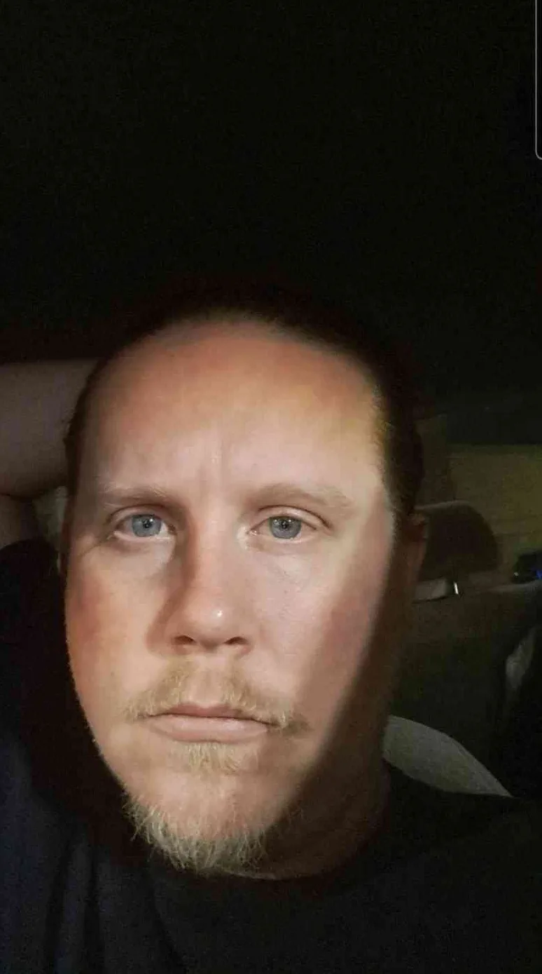 A close-up selfie of a man with light-colored eyes and a light beard, looking directly at the camera with a tired…
