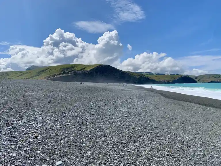 A wide-angle photo of a vast, pebbly black beach under a bright blue sky with large white clouds. Turquoise ocean waves…