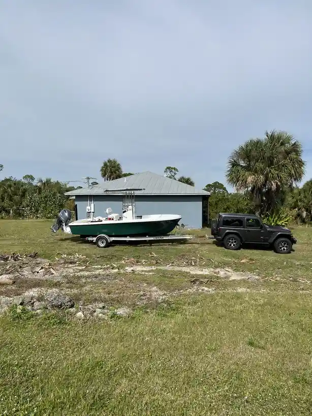 A dark gray Jeep Wrangler is parked on grass, hitched to a trailer carrying a green and white fishing boat. Behind them is a…