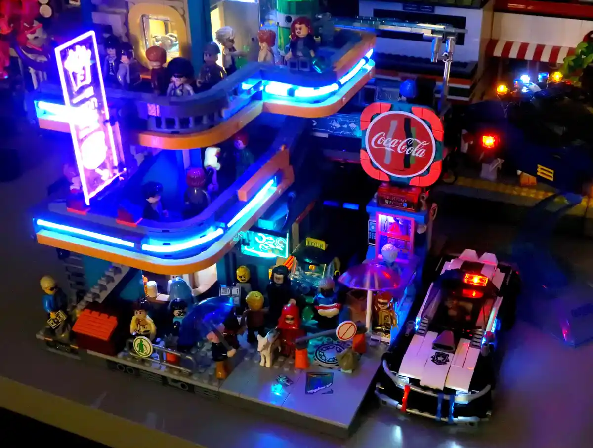 A detailed, illuminated LEGO city scene at night, featuring a multi-level building with glowing blue neon lights, a…