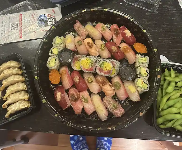 A top-down photo shows a large black takeout container filled with assorted sushi, including nigiri and maki rolls…