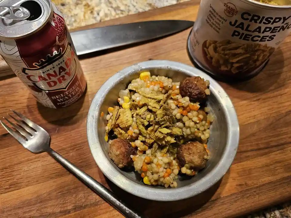 A metal bowl of couscous mixed with meatballs and topped with crispy jalapeño pieces sits on a wooden cutting board next to…