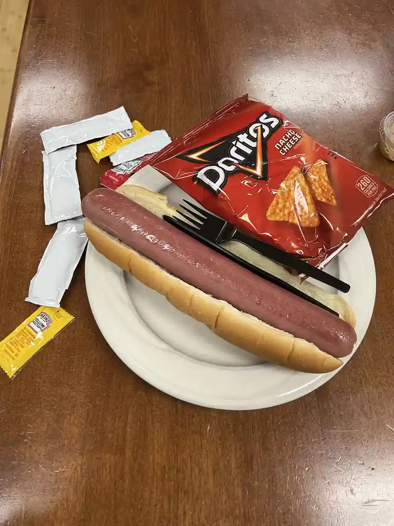 A plain hot dog in a bun sits on a white plate with a black fork and knife, next to a bag of Doritos Nacho Cheese chips and…