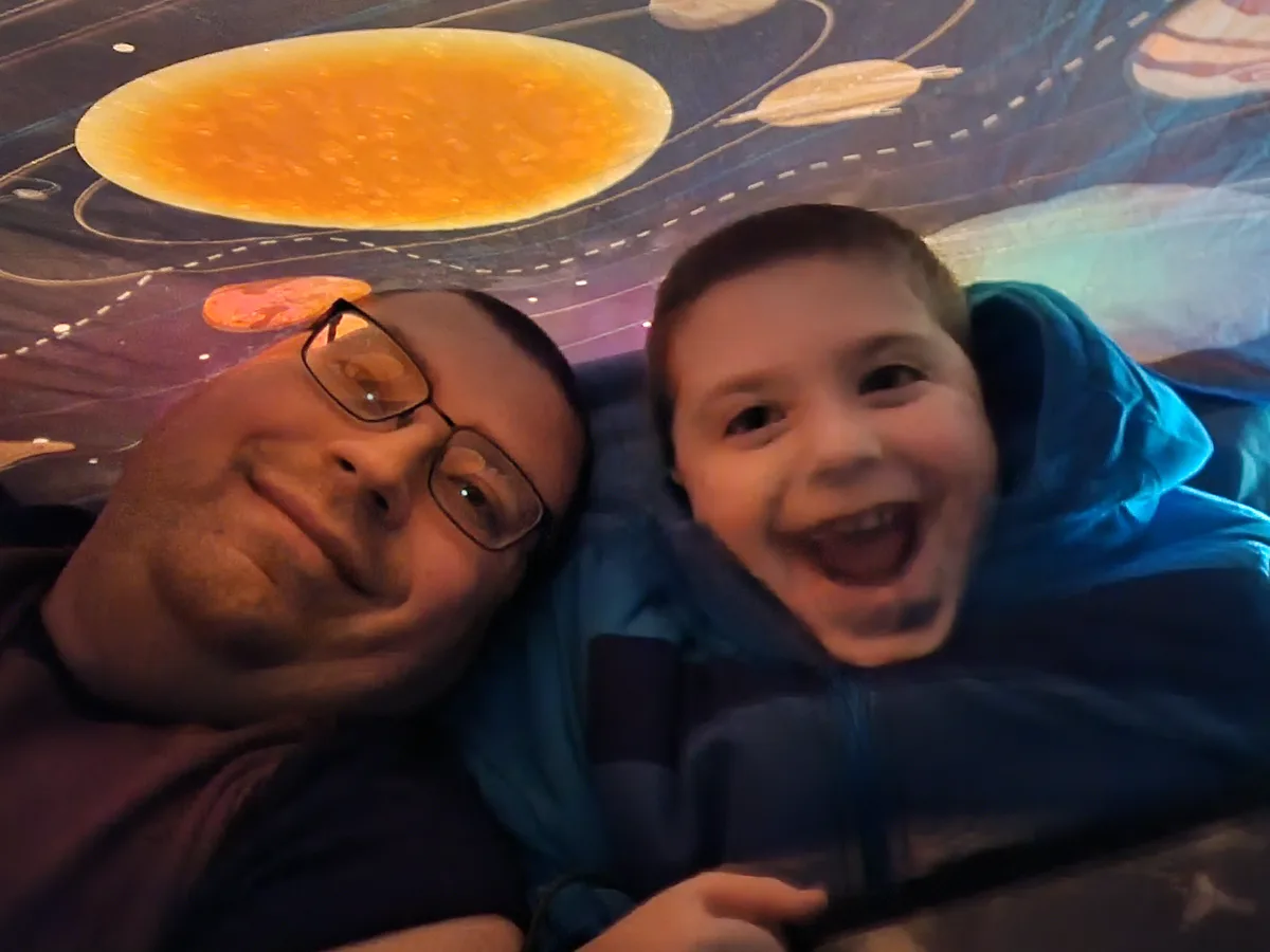A man and a young boy lie side-by-side on a bed with a space-themed blanket featuring planets and orbits. The man, wearing…