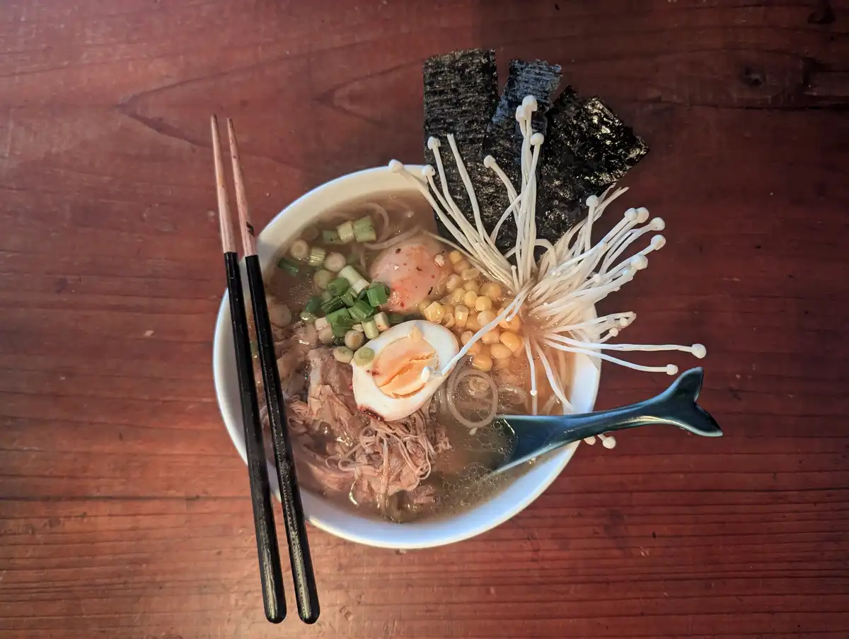 a top-down photo of a white bowl of ramen on a wooden table, containing broth, noodles, sliced pork, a soft-boiled egg…