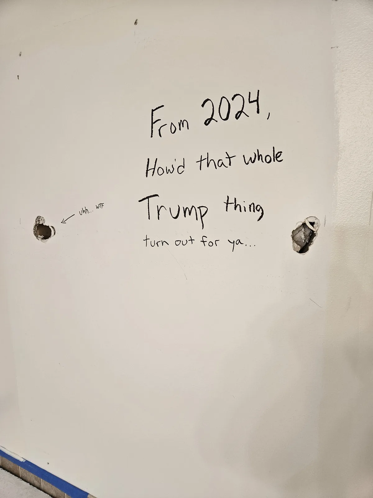 A white wall with two holes punched in it, one on the left and one on the right. Handwritten text in black marker reads…