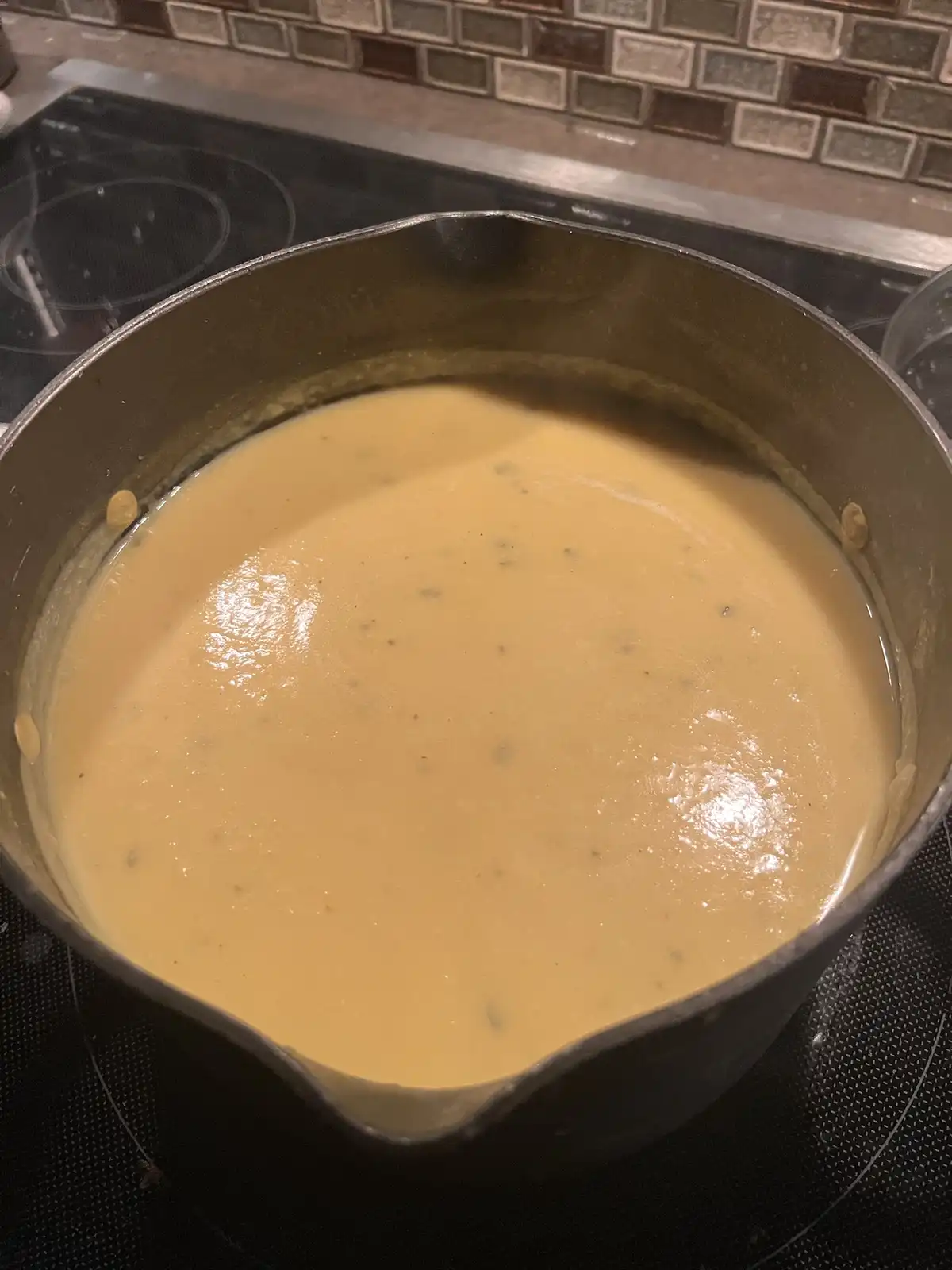 A close-up photo shows a large metal saucepan on a black electric stovetop, filled with a smooth, creamy, pale yellow sauce…