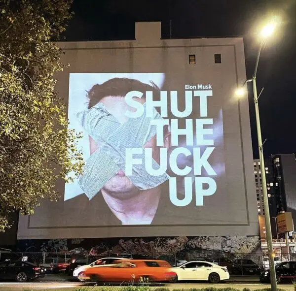 A large outdoor billboard at night shows a photo of Elon Musk with his mouth covered by duct tape. Bold white text overlaid…