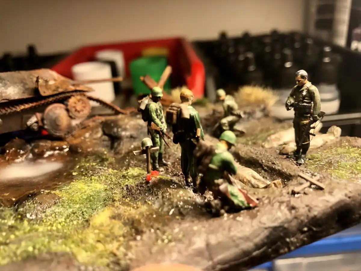 A diorama of miniature World War II soldiers in a muddy battlefield scene, with one soldier kneeling beside a makeshift…