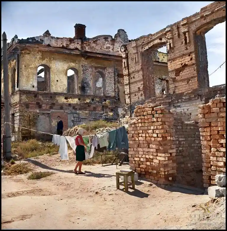 A woman in a red shirt and green skirt hangs laundry on a line in front of a ruined brick building with broken windows and…