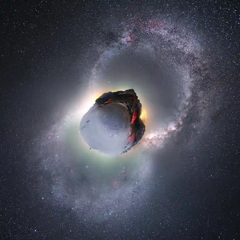 A 360-degree panoramic image shows a snowy mountain peak surrounded by the Milky Way galaxy, with a small human figure…