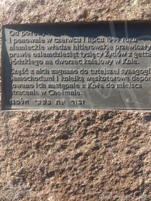 A close-up photograph of a dark metal plaque mounted on a rough, textured stone wall. The plaque contains engraved text in…