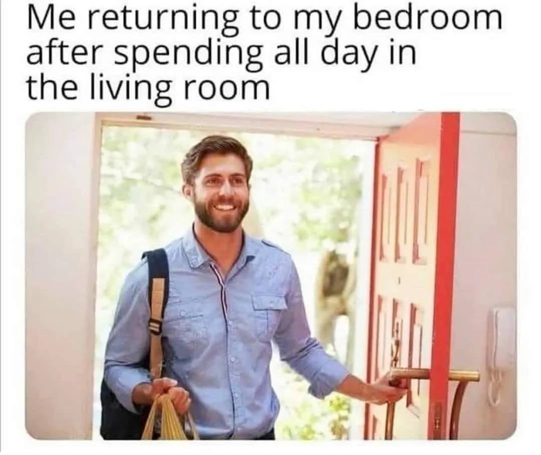 A man with a beard and blue shirt smiles brightly while entering a room through a red door, holding a bag and a bundle of…
