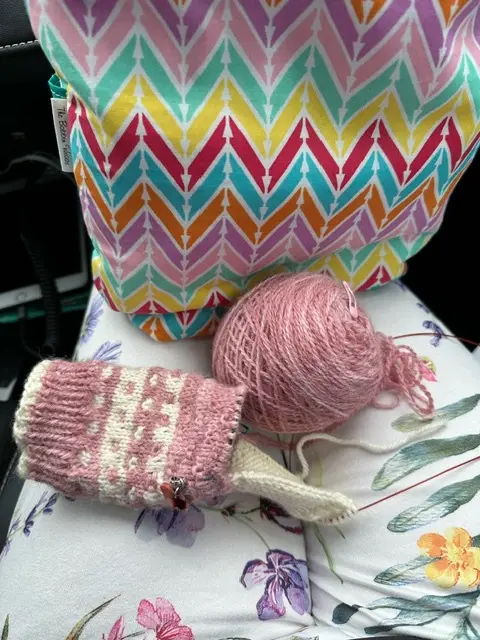 A close-up photo shows a partially knitted pink and cream patterned sock lying next to a ball of pink yarn, both resting on…