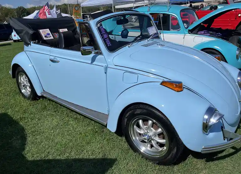 A light blue Volkswagen Beetle convertible is parked on grass at a car show, with its top down and chrome wheels visible…